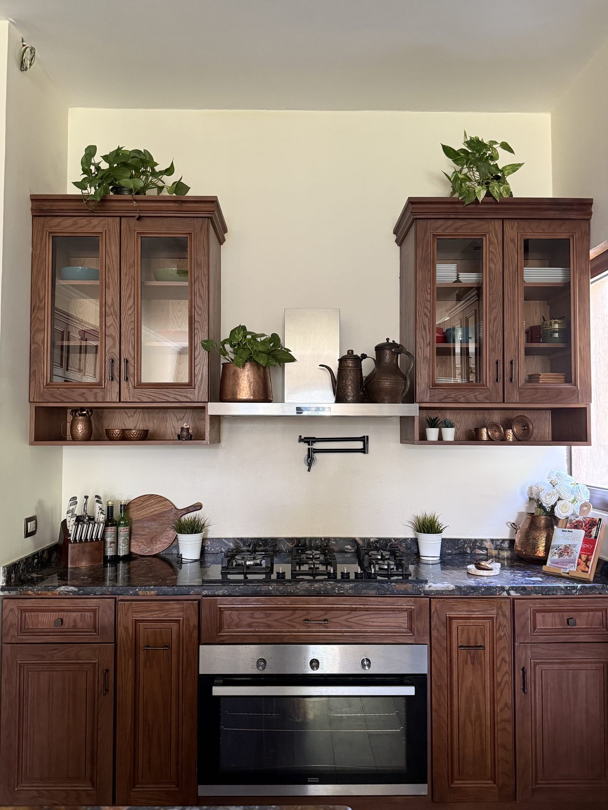 The first D’s kitchen — glass-front cabinets flanking the gas range, in warm wood with dark granite worktops