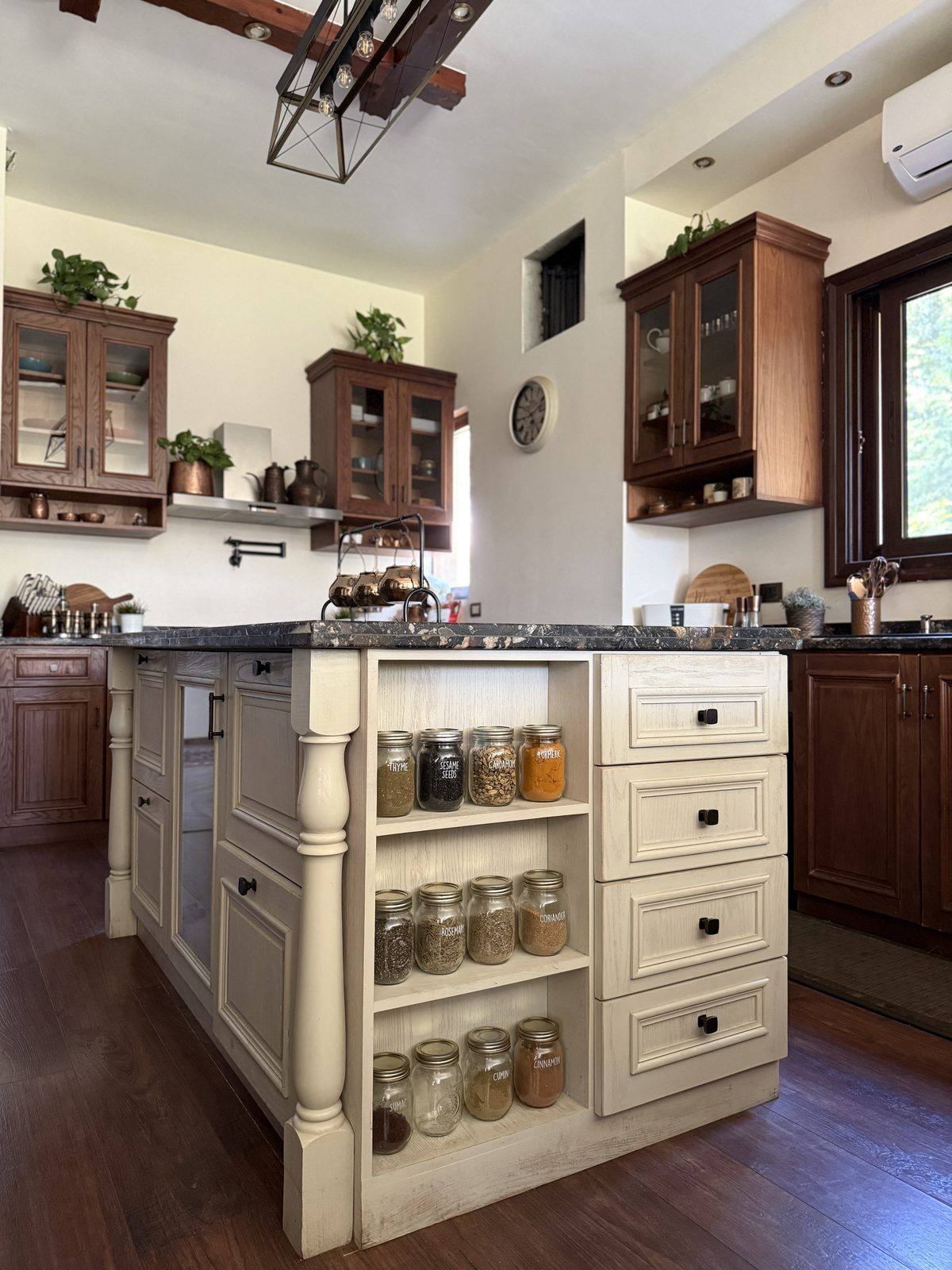 A cream painted island with turned legs and open spice shelving, against warm wood cabinetry